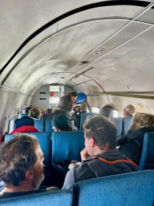 Passengers sit in blue seats inside a small aircraft, some wearing winter clothing, likely for an overnight trip at the South Pole. A flight attendant stands near the front, attending to a passenger. The cabin is narrow with a curved white ceiling.