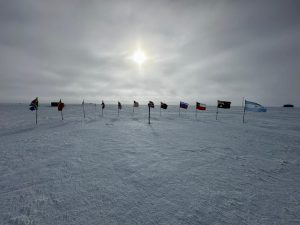 A row of international flags stands in the snow under a cloudy sky with the sun shining dimly overhead, marking the start of an unforgettable overnight trip at the South Pole in this vast, icy landscape.