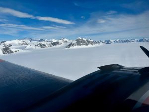 A view from an airplane window shows snow-covered mountains and a vast, icy landscape beneath a blue sky—an unforgettable scene during an overnight trip at the South Pole, with the aircraft wing and engine visible in the foreground.