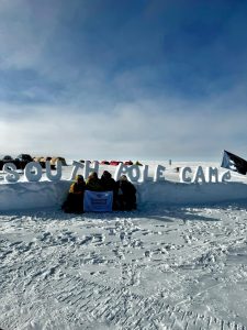 A group of people sit on snow in front of large letters spelling “SOUTH POLE CAMP,” bundled in heavy winter gear after an unforgettable overnight trip at the South Pole, with tents, a flag, and blue sky in the background as they hold a sign.
