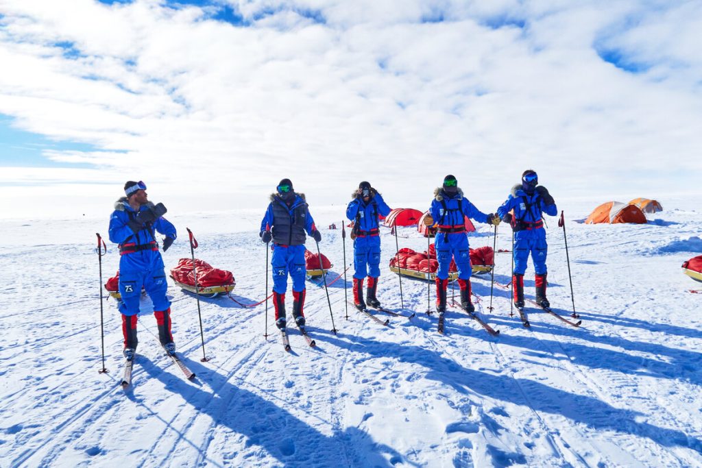 Five people in blue and red snowsuits stand on skis in a snowy landscape, each with a sled behind them. Orange tents are set up in the background under a partly cloudy sky.
