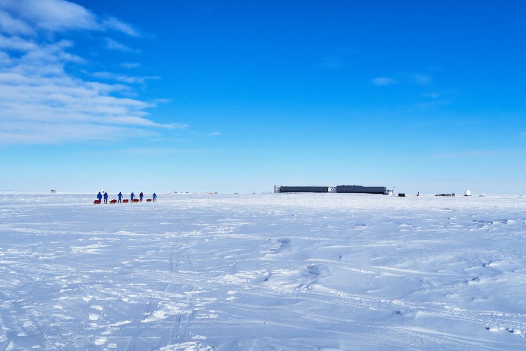 A group of people pulling sleds across a vast, snowy landscape under a blue sky, with a large, dark building and smaller structures in the distance.