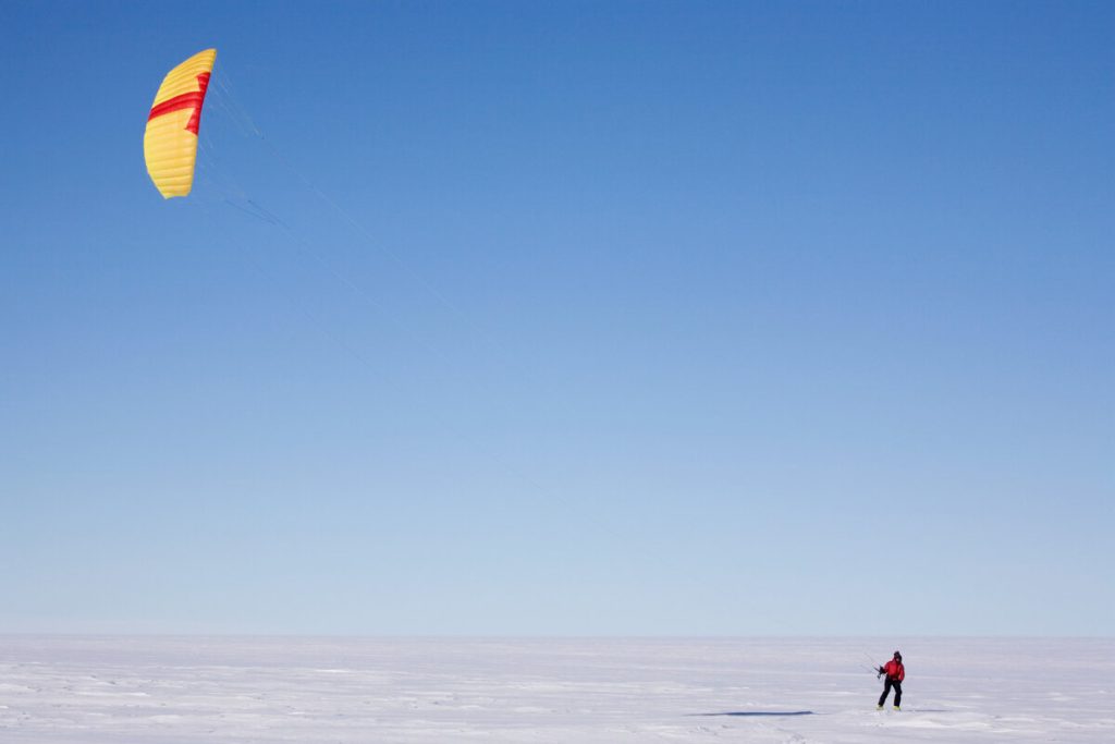 A person in winter clothing stands on a snowy, flat landscape while flying a large yellow and red kite under a clear blue sky.