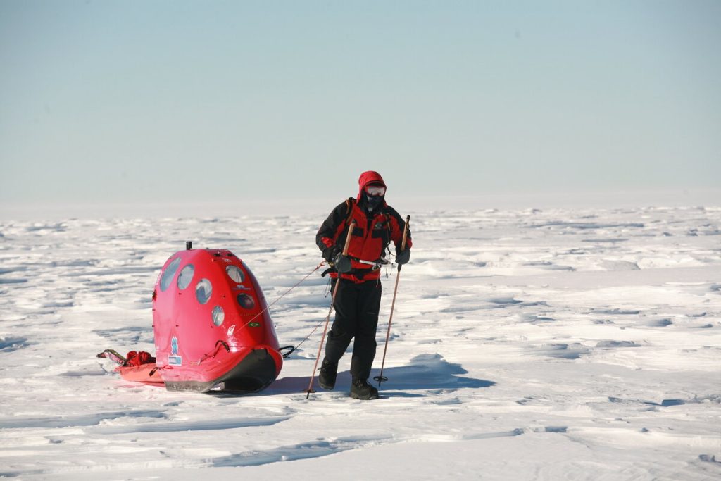 A person in cold-weather gear pulls a red sled with windows across a vast, snowy, and icy landscape under a clear sky.
