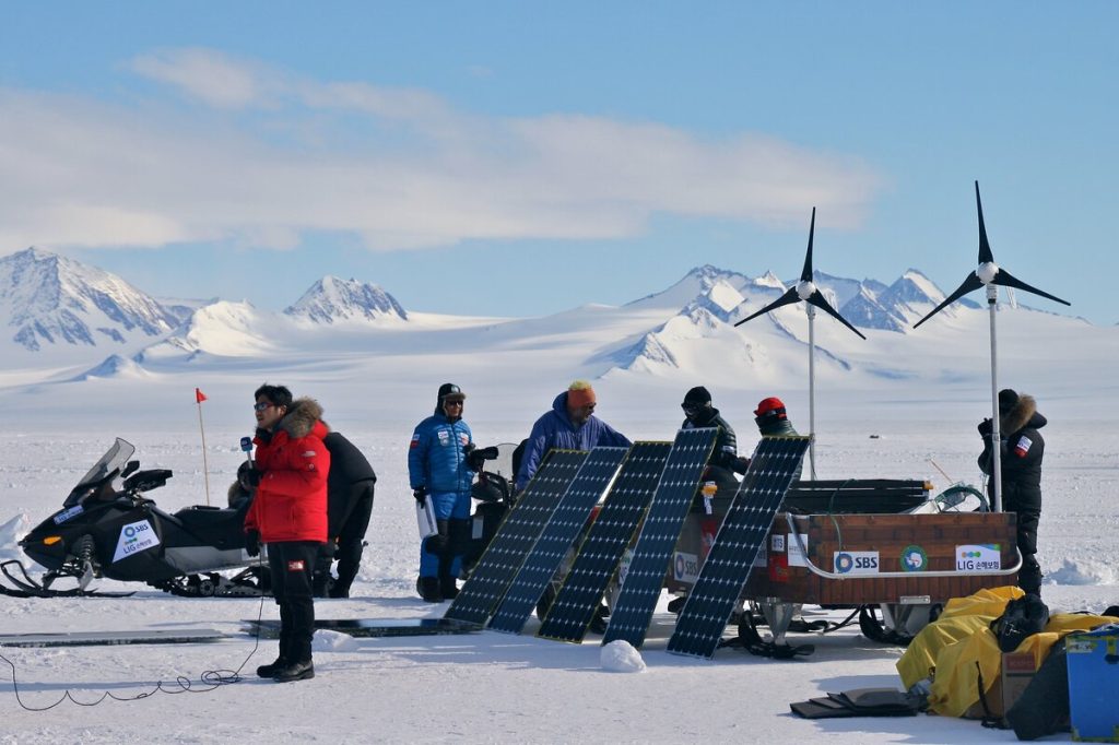 A group of people in winter gear work with solar panels and wind turbines on a snowy landscape with mountains in the background; snowmobiles and equipment are nearby.