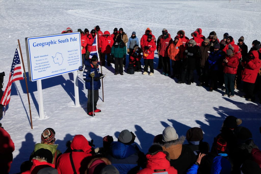 A group of people in winter gear gather around a speaker at the Geographic South Pole marker, with a large informational sign and the U.S. flag visible on a snow-covered landscape.