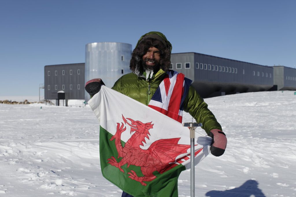 A person in cold weather gear stands on snow holding a Welsh flag, with a Union Jack draped over their shoulder. A modern, rectangular building is in the background under a clear blue sky.