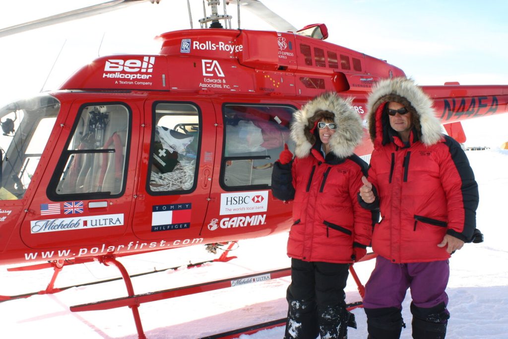 Two people in red parkas with fur-lined hoods stand in front of a red helicopter on snow. They are wearing sunglasses and smiling, with one giving a thumbs-up. The helicopter has various sponsor logos and texts on it.