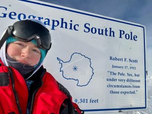 A person in cold-weather gear and goggles takes a selfie during an overnight trip at the South Pole, standing in front of a sign reading Geographic South Pole with a map of Antarctica and a quote from Robert F. Scott dated January 17, 1912.